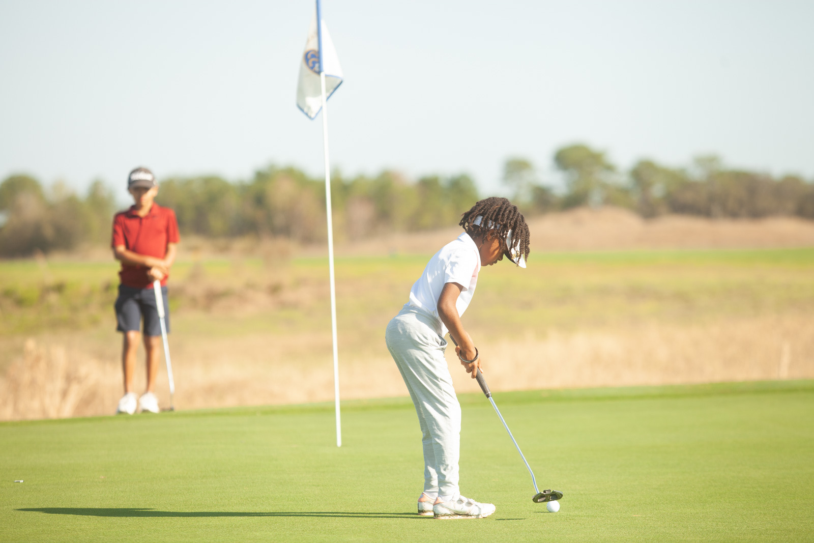 A junior golfer putting toward the hole while another young golfer watches